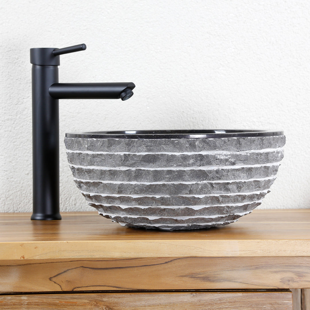 Round countertop washbasin made of natural stone with a structured surface on a wooden top, combined with a black stand-alone tap