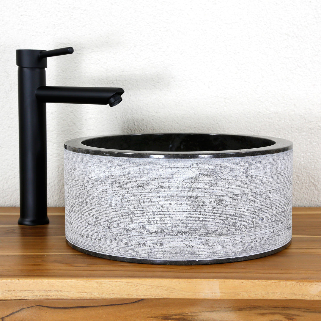 Round countertop washbasin made of natural stone with a striking line structure, combined with a black stand-mounted tap on wood.