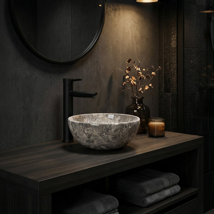 Round natural stone countertop washbasin with grey-brown grain on a dark wooden console in the atmospherically lit bathroom, combined with black fittings, mirror and subtle accessories
