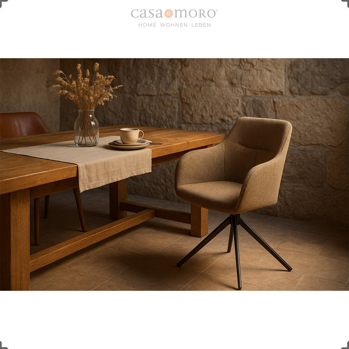 360° rotating upholstered chair with armrests in brown at a wooden table in a dining room with a natural stone wall and warm lighting ambience