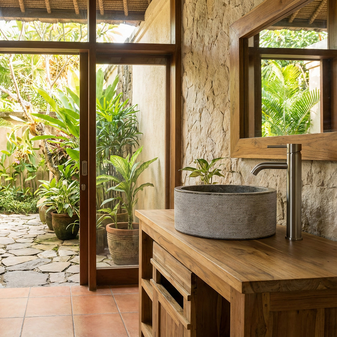 Round stone washbasin in gray stone on a wooden vanity unit, placed in the light-flooded bathroom with a garden view.