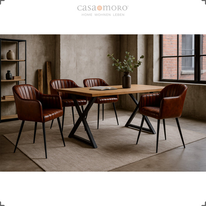 Four antique brown leather armchairs with vertical quilting at a wooden table with a black metal frame in a modern loft dining room with a concrete wall and large windows.
