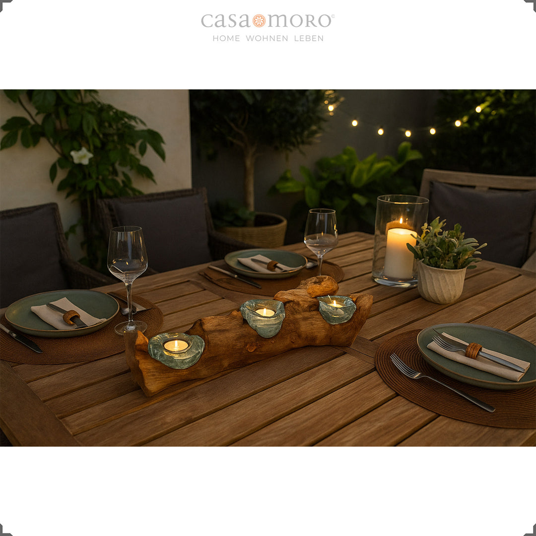 Three-part tea light holder made of glass and root wood as an atmospheric table decoration on a terrace, framed by plants and a set table