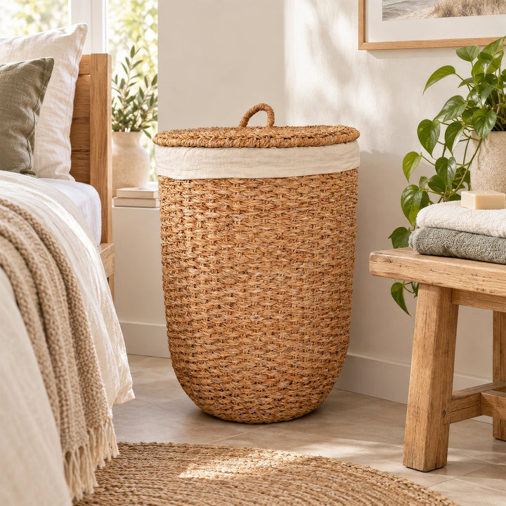 A laundry basket with a lid made of natural-colored seagrass and a light-colored fabric insert, placed between the bed and the wooden bench in the bedroom.
