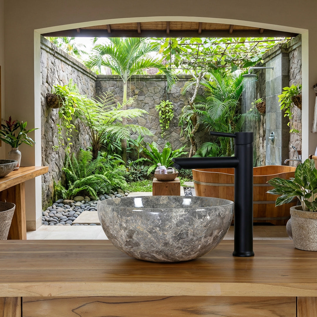 Round natural stone Washbasin with gray-brown grain on a wooden washbasin in the open spa bathroom with a view of a green courtyard, combined with a black stand-alone fitting, natural stone wall and wooden bathtub
