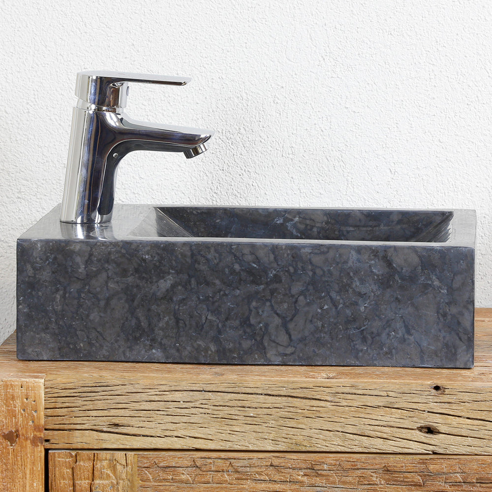 Square countertop basin made of marble with dark grain on a wooden console, combined with chrome-plated fittings in front of a light wall